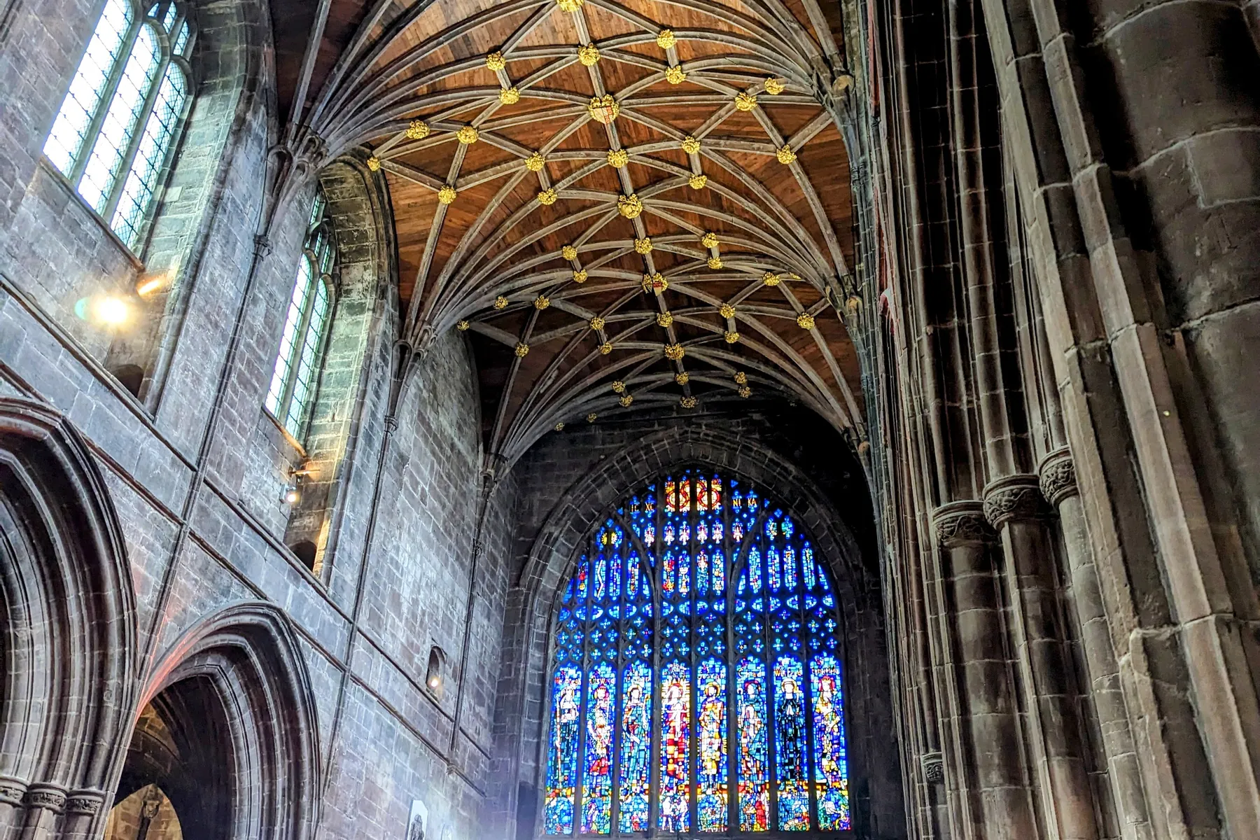 The vaulted wooden ceiling of Chester Cathedral, studded with gold, above sandstone arches and a tall stained glass window