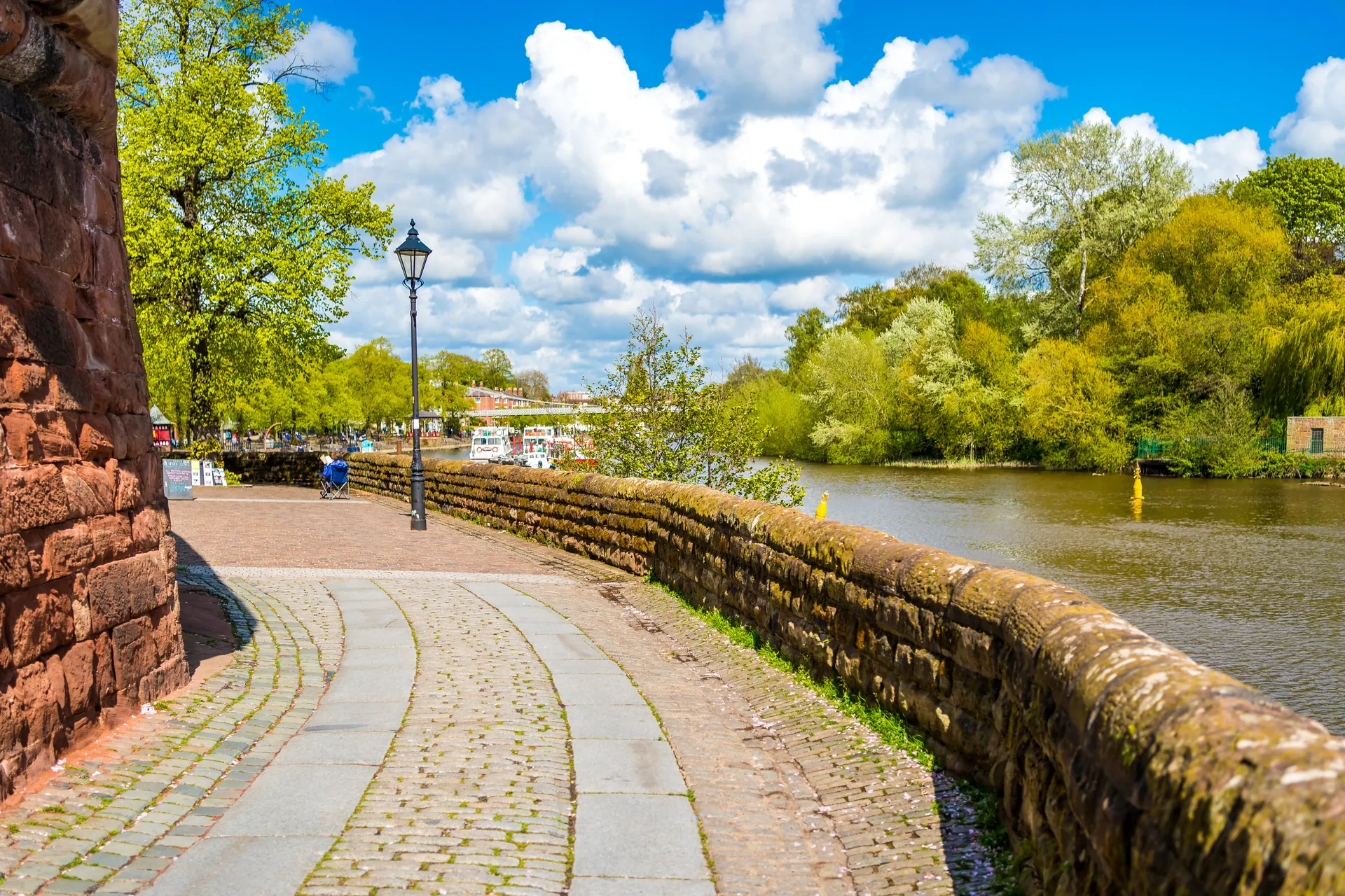 The city walls walk curving beside the River Dee, with an old lamp post and green trees under a bright sky