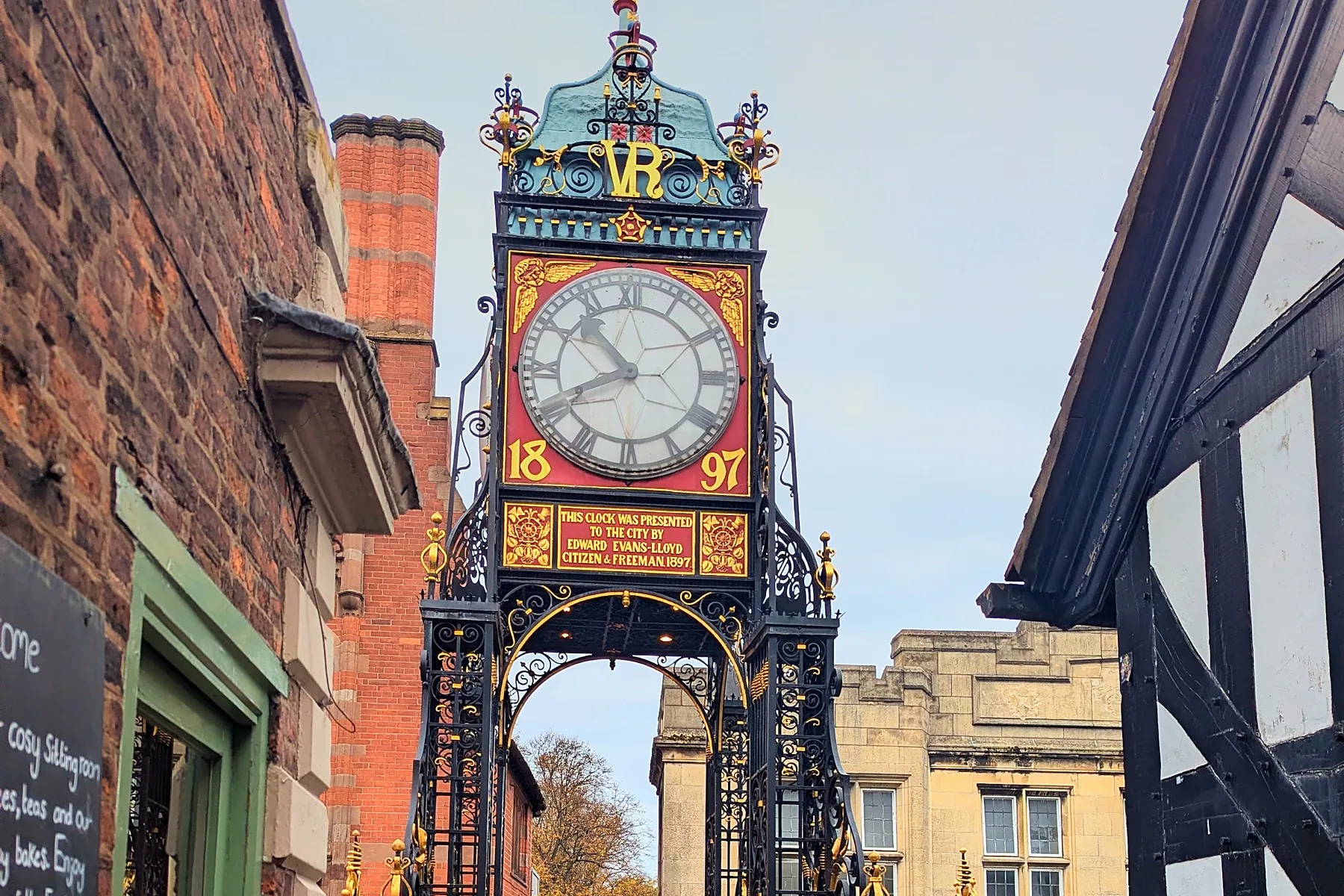 The Eastgate Clock seen along the wall walk, its red, blue and gold ironwork between green railings and timber framed buildings