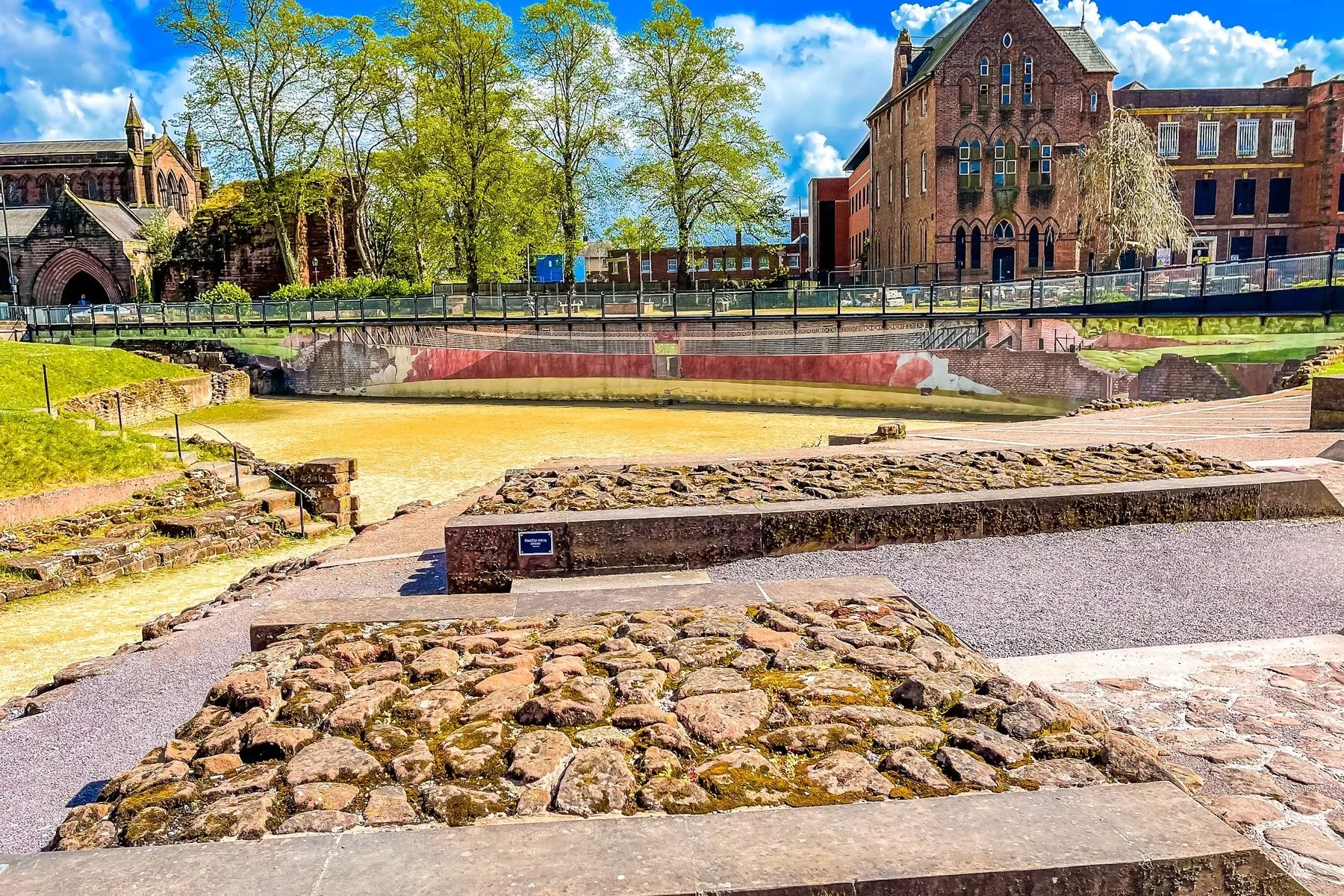 The low stone walls and sandy arena of the Roman Amphitheatre in Chester under a bright blue sky