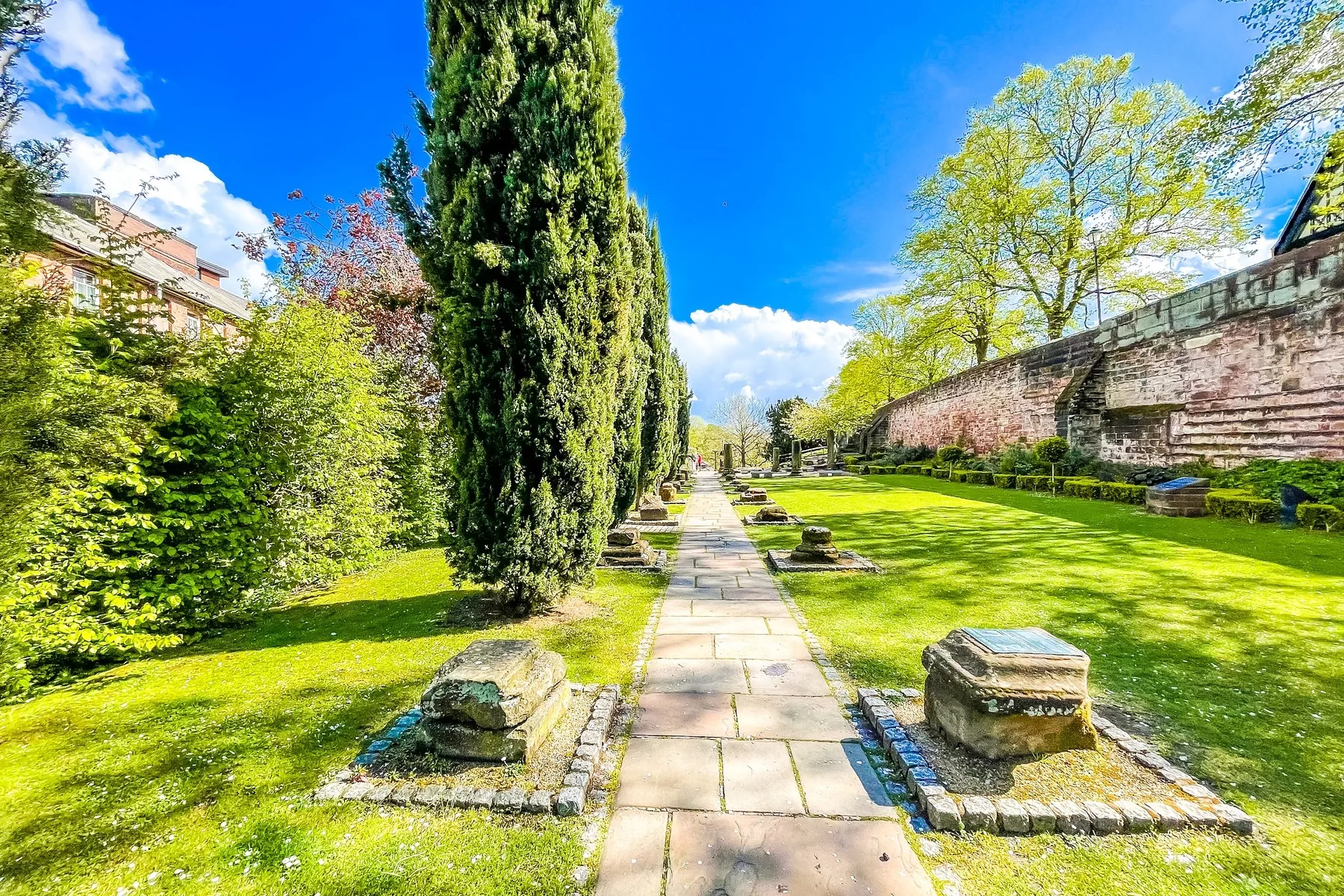 A stone path between green lawns and tall cypress trees in Chester's Roman Gardens, with the red sandstone city walls on the right