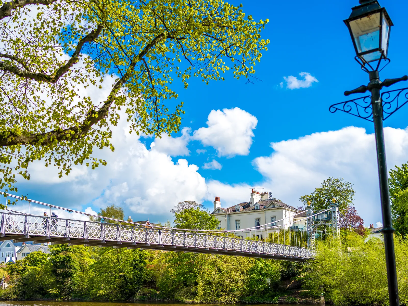 The Queen's Park suspension bridge over the River Dee, seen through green trees beside a lamp post