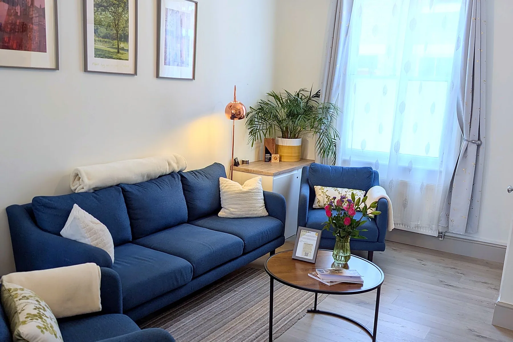 The lounge at Enjoy the Pause: blue sofas, a round coffee table with flowers, and light from the front window