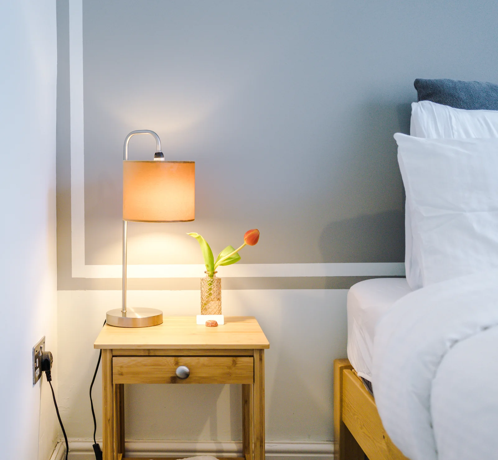 Bedside table with a warm lamp and fresh tulip beside the made-up bed